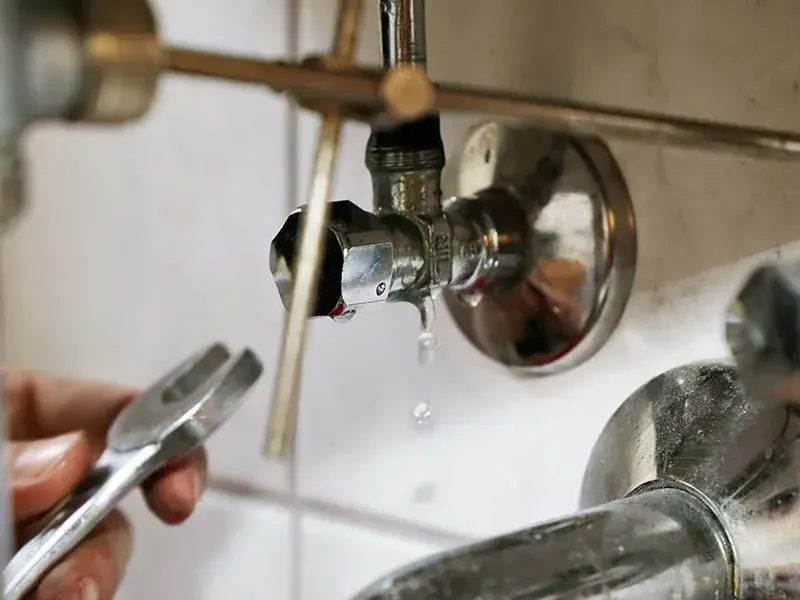 Water dripping from a chrome angle stop valve as a hand holds an open-end wrench nearby