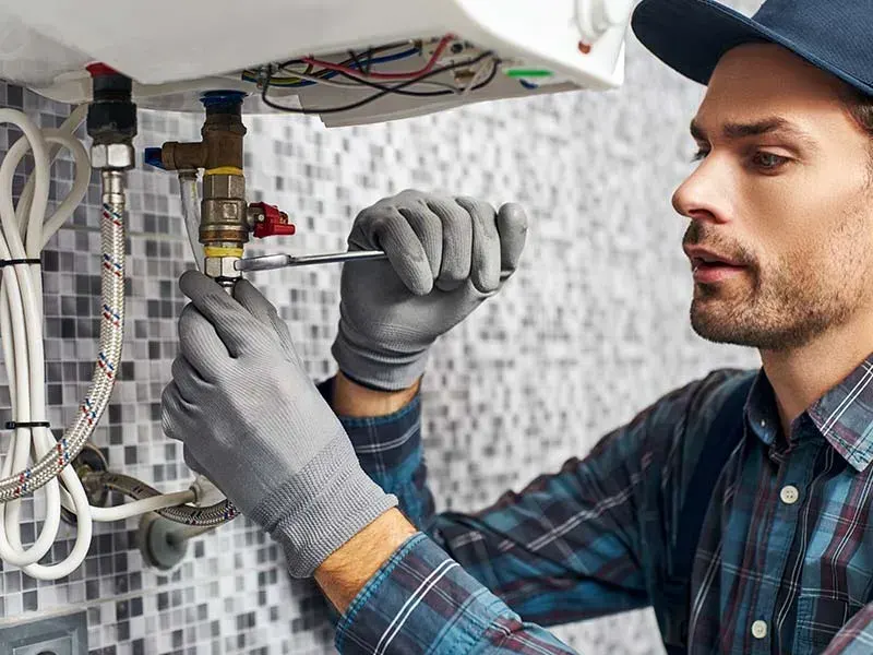 Plumber in gloves using a screwdriver on the valve fittings under a wall-mounted water heater
