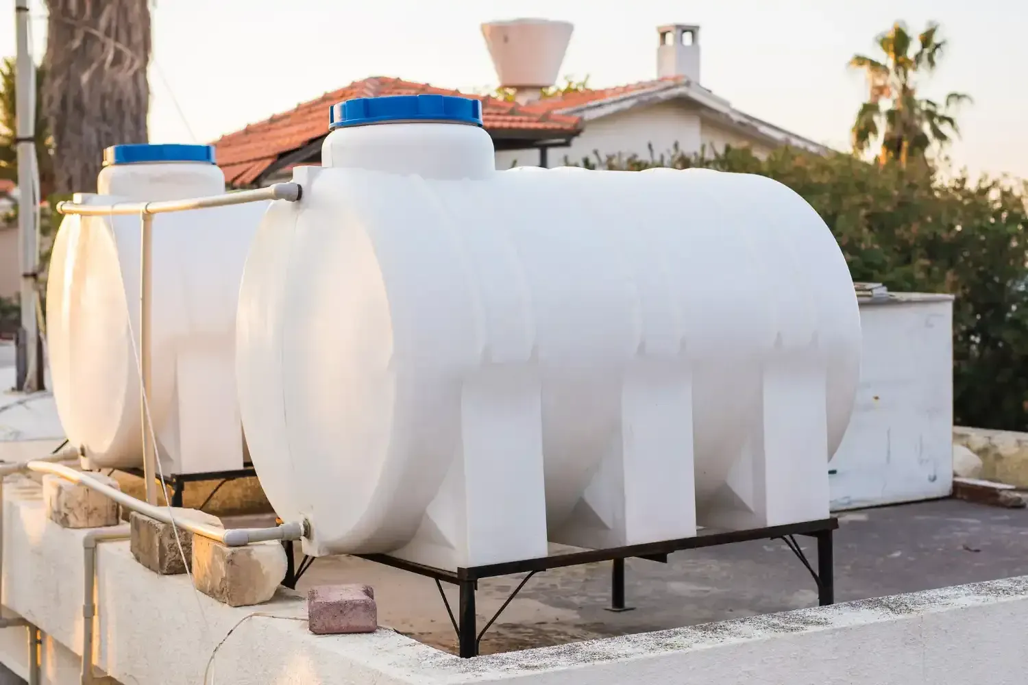 Two white plastic water storage tanks with blue caps on metal stands on a rooftop