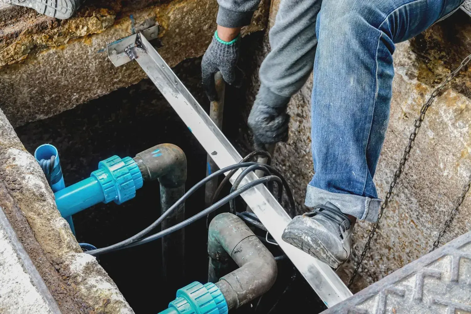 Worker in gloves climbing into an underground utility vault with blue PVC and gray water pipes